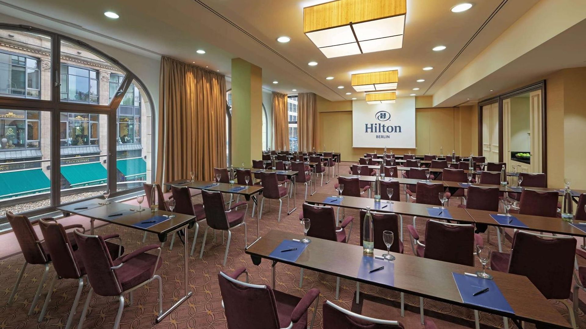 1548869662 Konferenzraum im Hilton Berlin mit Tischen, Stühlen und großen Fenstern, die Tageslicht hereinlassen.