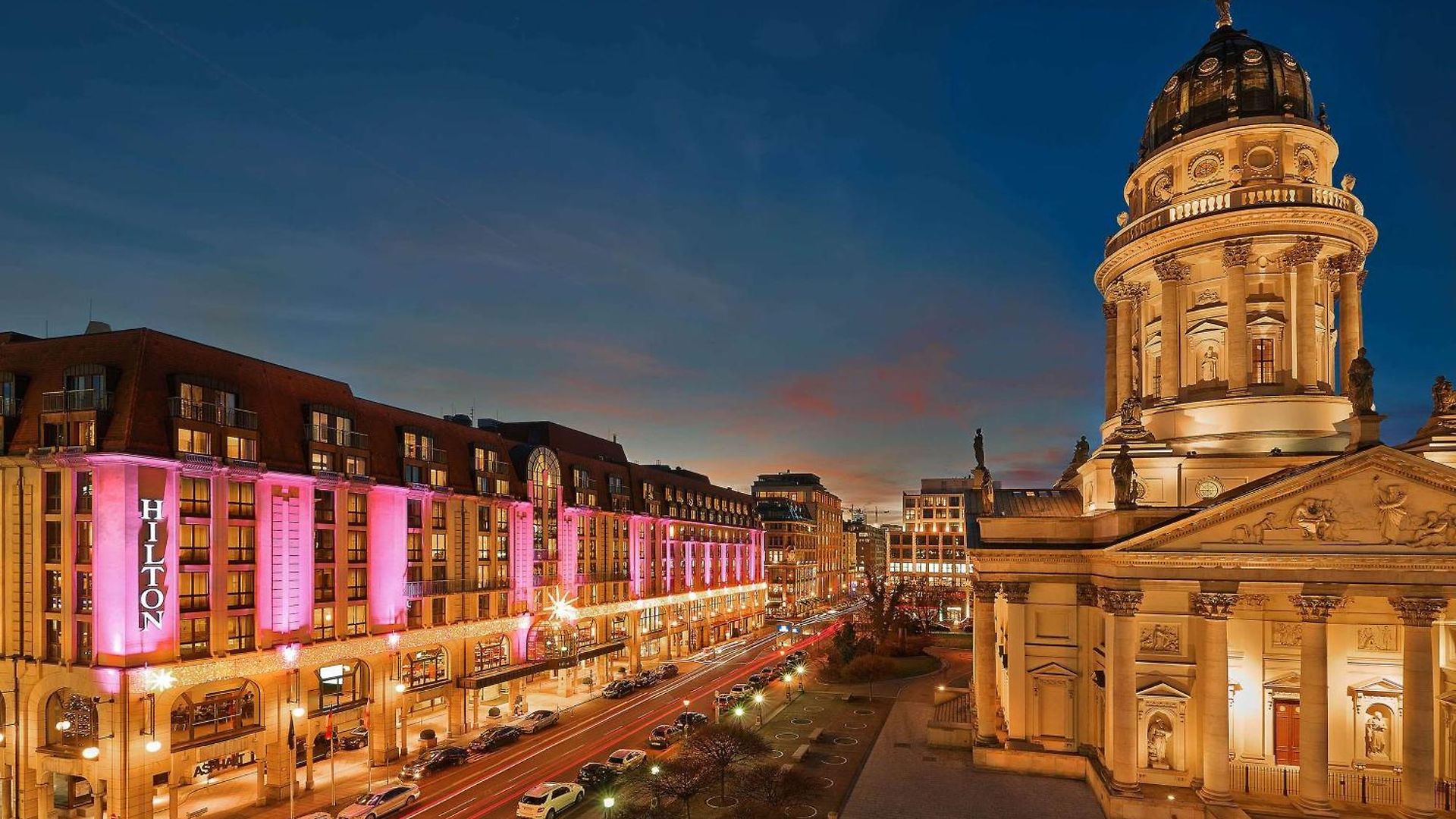 Blick auf das Hilton Hotel mit pinker Beleuchtung und einer historischen Kirche bei Dämmerung.