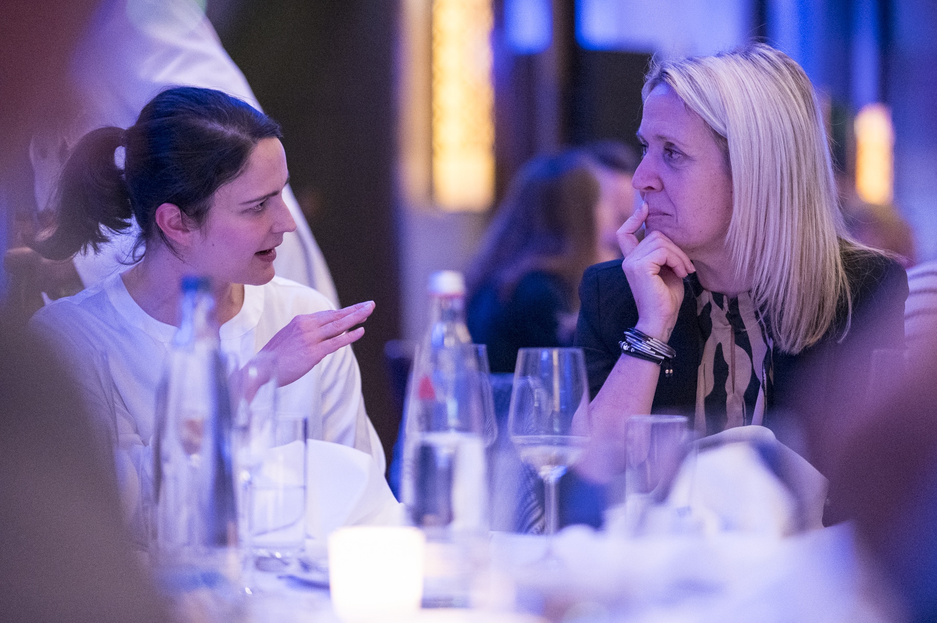 CSebastian Hoehn Zwei Frauen sitzen an einem Tisch, unterhalten sich lebhaft, umgeben von Gläsern und Kerzenlicht.