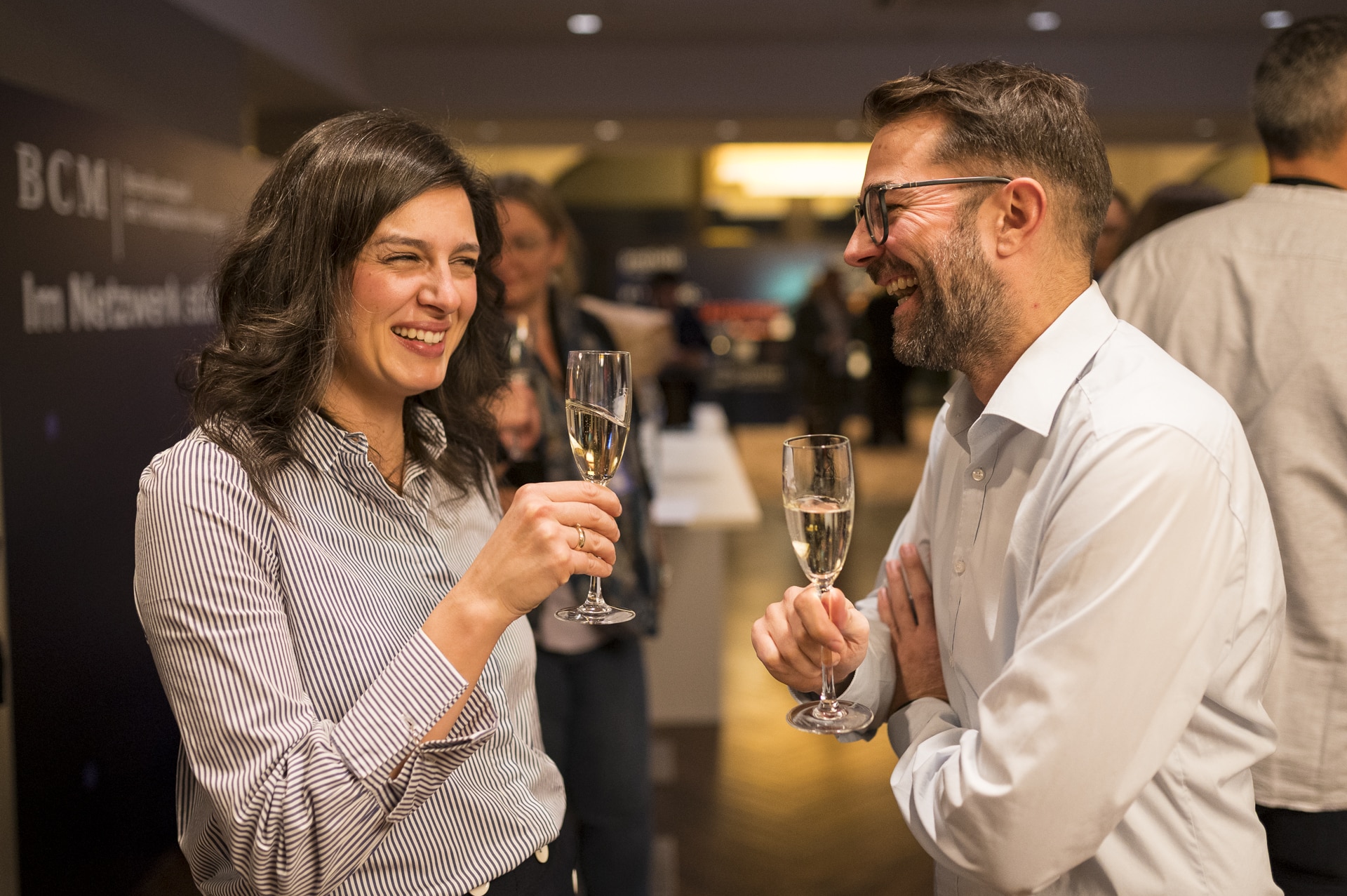 cSebastian Hoehn Zwei Personen halten Sektgläser, lächeln und unterhalten sich in einem festlichen Umfeld.