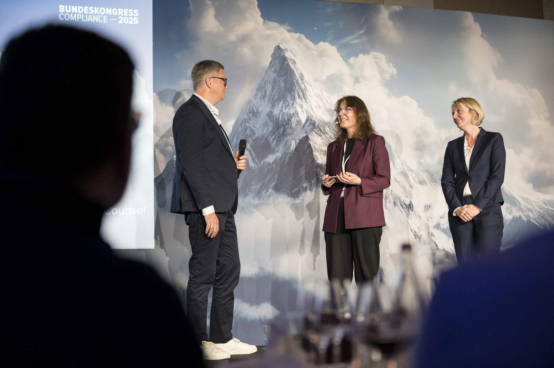 cSebastian Hoehn Drei Personen stehen auf einer Bühne vor einer Berglandschaft, während eine Frau einen Preis entgegennimmt.