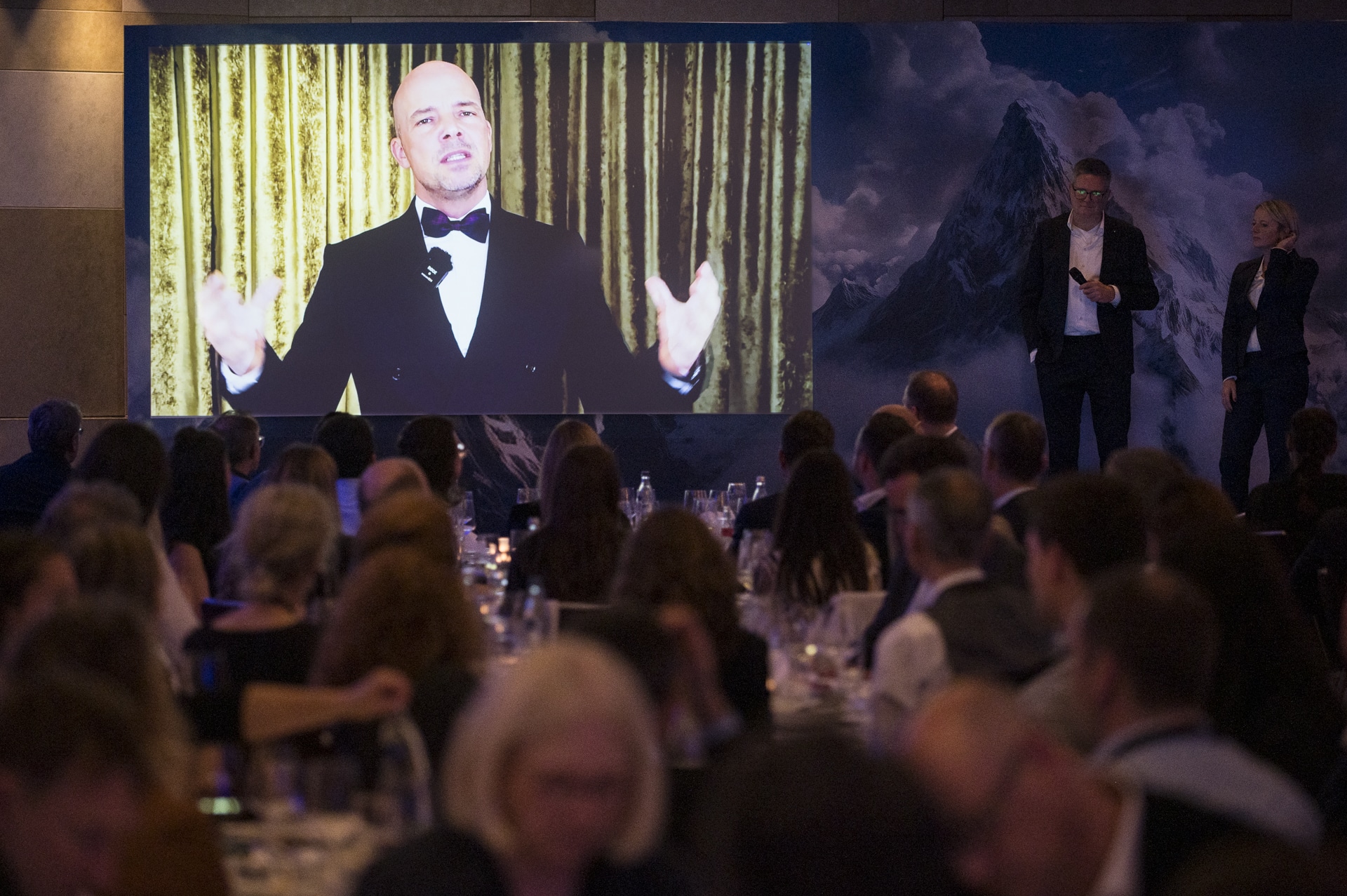 cSebastian Hoehn Mann in Smoking spricht auf großer Leinwand, während Publikum bei einer Veranstaltung aufmerksam zuhört.