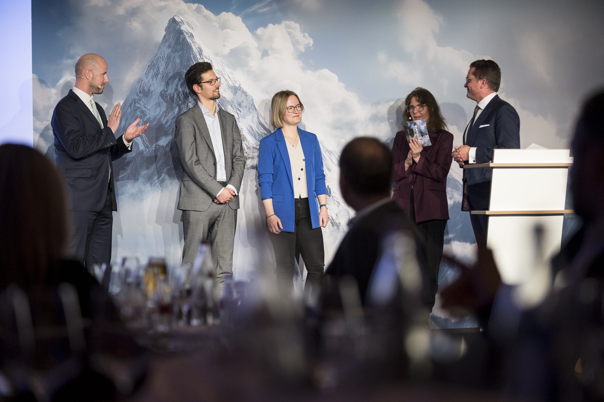 cSebastian Hoehn Fünf Personen stehen auf einer Bühne vor einer Berglandschaft, während sie sich gegenseitig applaudieren und sprechen.