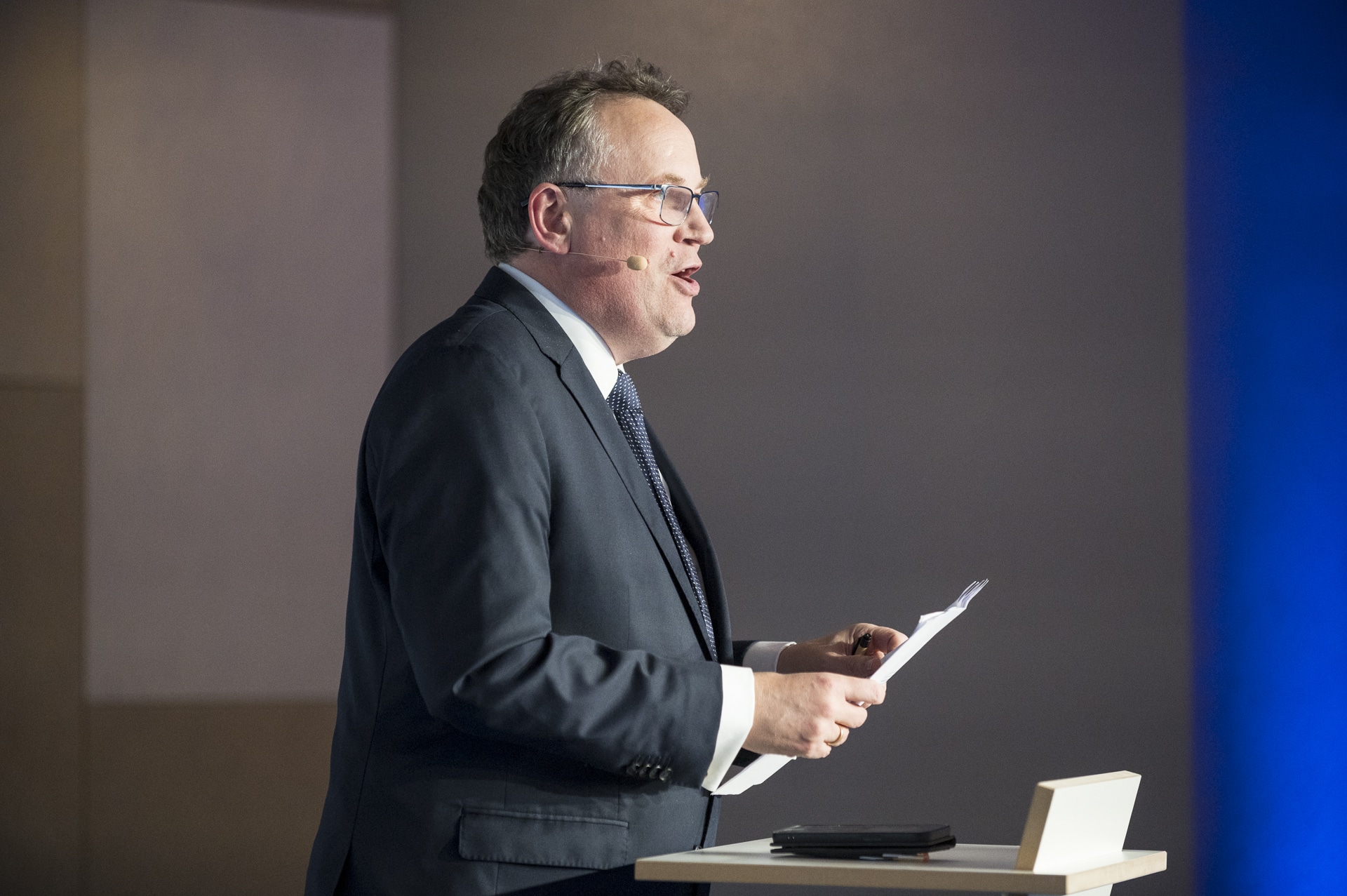 cSebastian Hoehn Mann in Anzug spricht am Rednerpult, hält ein Blatt Papier, mit Mikrofon am Ohr, im Hintergrund unscharfe blaue Wand.