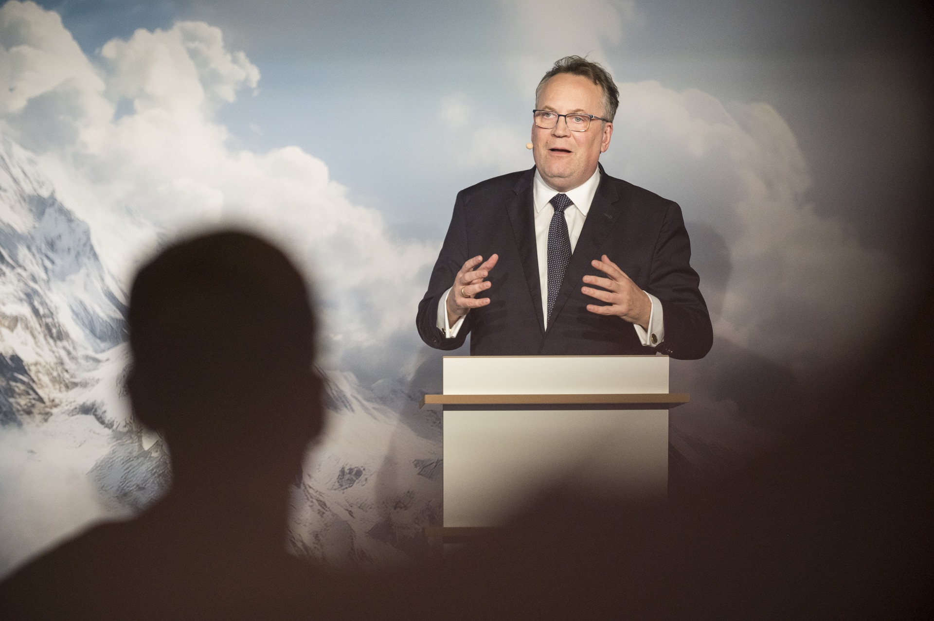 cSebastian Hoehn Mann in Anzug spricht an einem Rednerpult vor einer bergigen, wolkigen Kulisse.