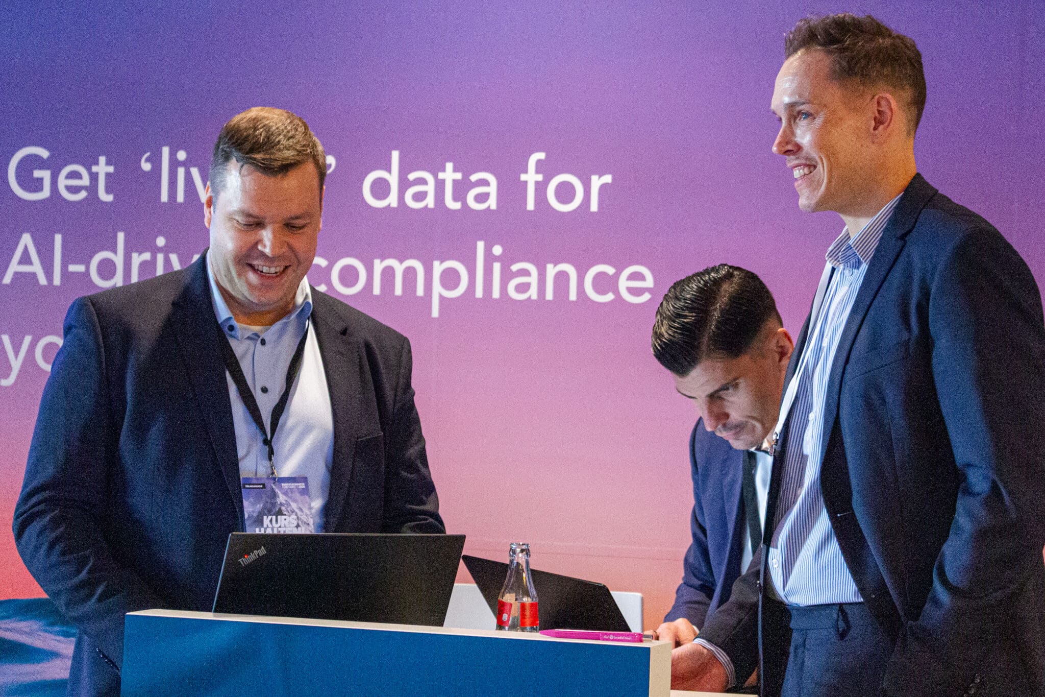 feldmann_robert_8583 Drei Männer stehen an einem Tisch, lächeln und diskutieren, während im Hintergrund ein Banner mit Text zu sehen ist.