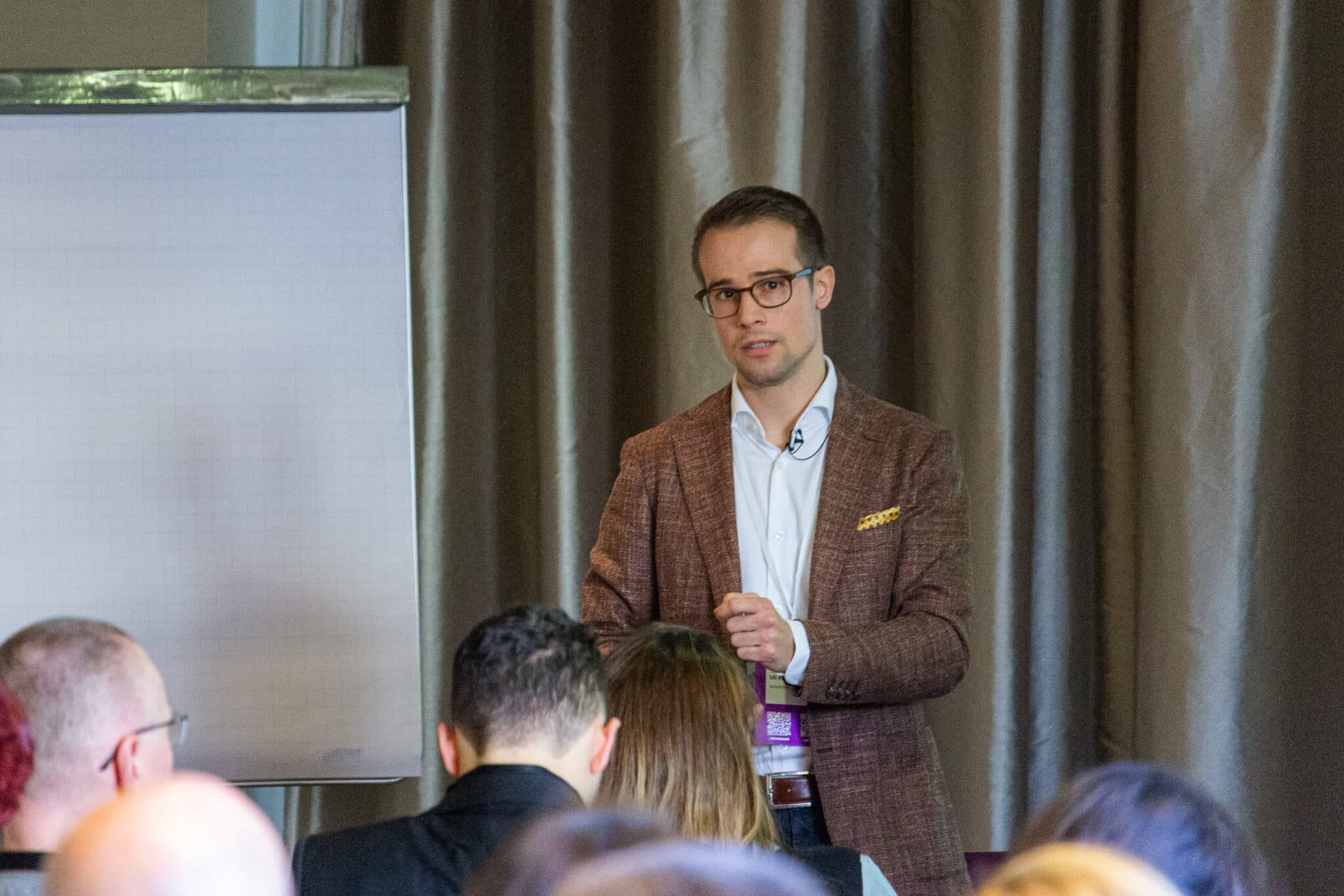 feldmann_robert_8612 Mann in Anzug spricht vor Publikum, mit Flipchart im Hintergrund und Vorhängen, die den Raum abgrenzen.
