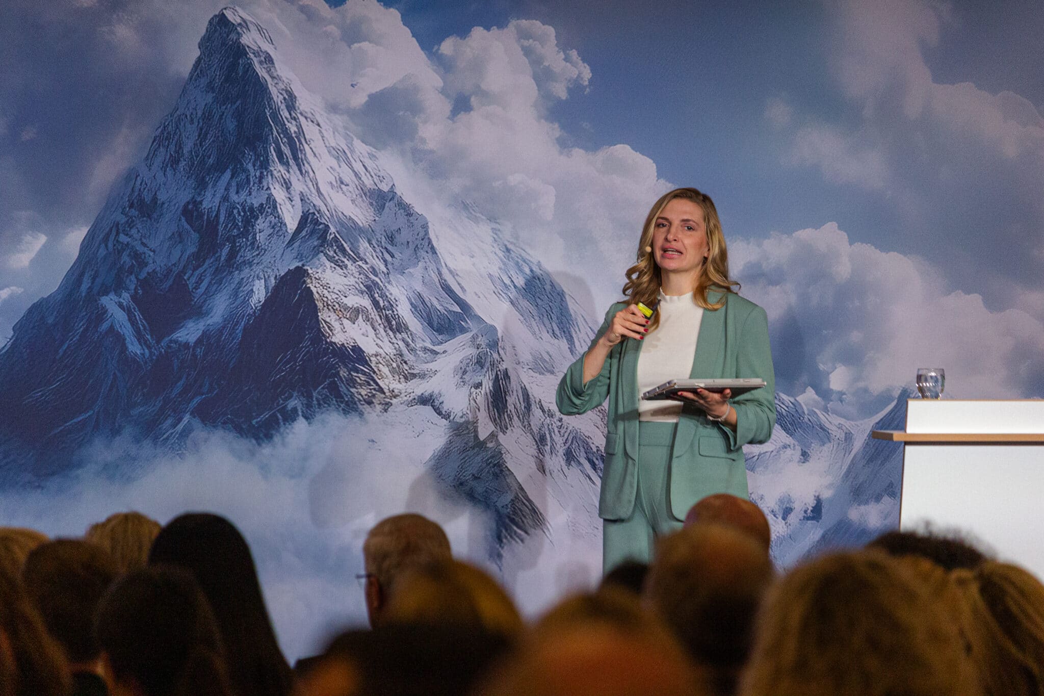 feldmann_robert_8676 Frau in hellgrüner Kleidung spricht vor einer Berglandschaft, hält ein Tablet und ein Mikrofon, Publikum im Hintergrund.