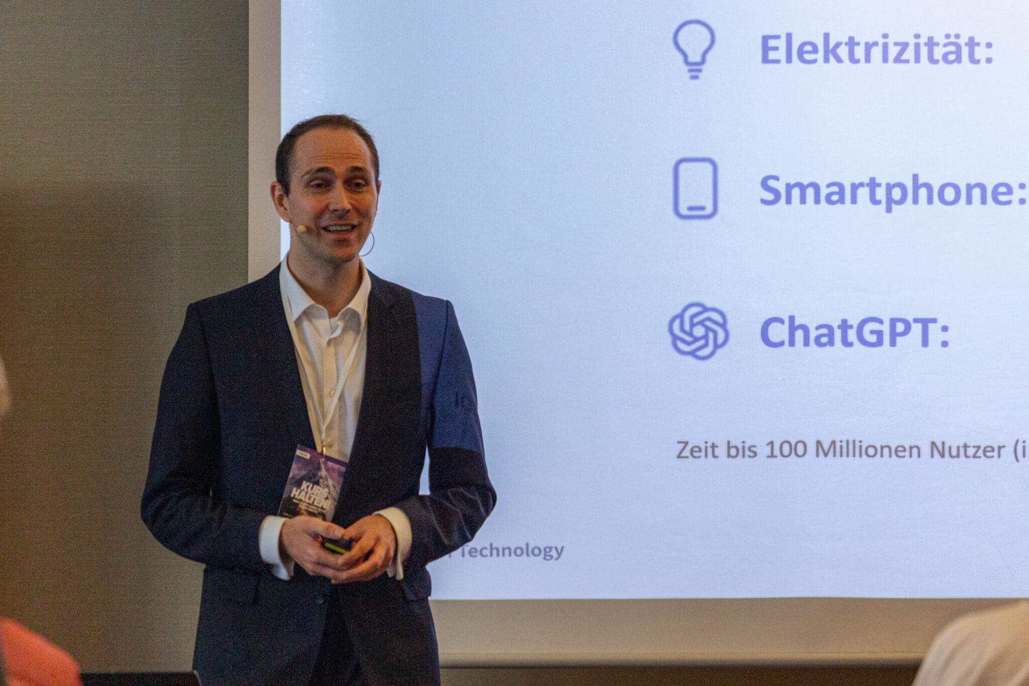 feldmann_robert_8710 Mann in Anzug spricht vor einer Präsentation mit Grafiken zu Technologie und Nutzung von ChatGPT.