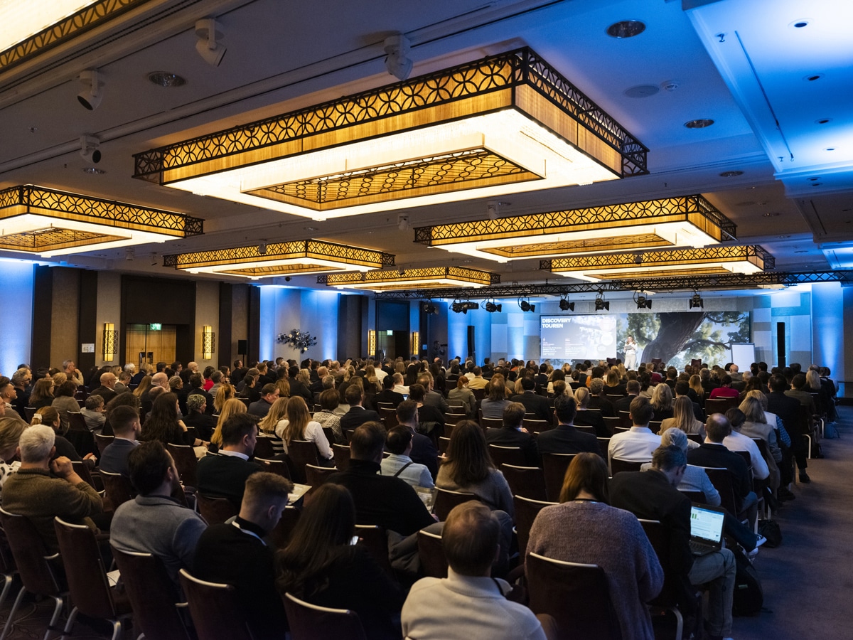 _DSF0707_1200x900 Large conference hall filled with attendees facing a stage and presentation screen under ornate hanging lights.